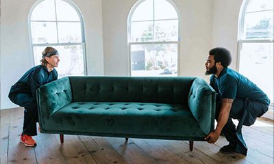 Two professional movers carefully lifting a green upholstered sofa in a bright room with large