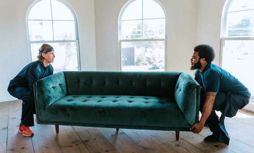 Two professional movers carefully lifting a teal sofa in a bright, spacious room with large