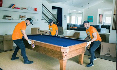 Skilled removalists in orange shirts carefully relocating a pool table in a modern home, showcasing