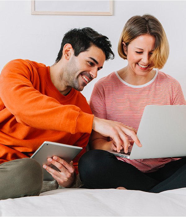Couple sitting together, smiling and discussing while using a laptop and tablet, representing the ease