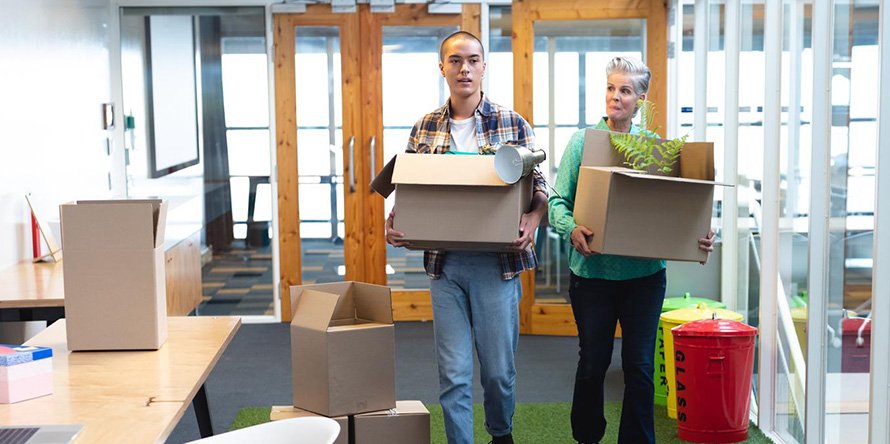 Two individuals carrying moving boxes and a plant in an office setting, illustrating the office