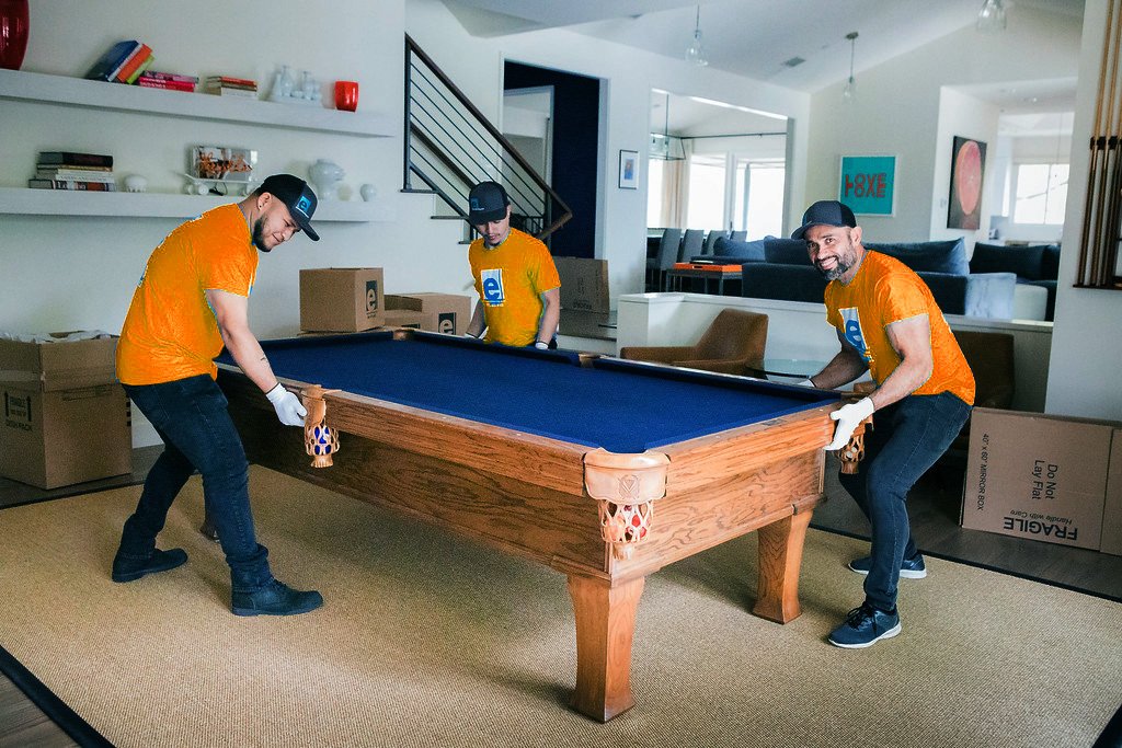 Two professional movers carefully lifting a pool table in a spacious living room, surrounded by